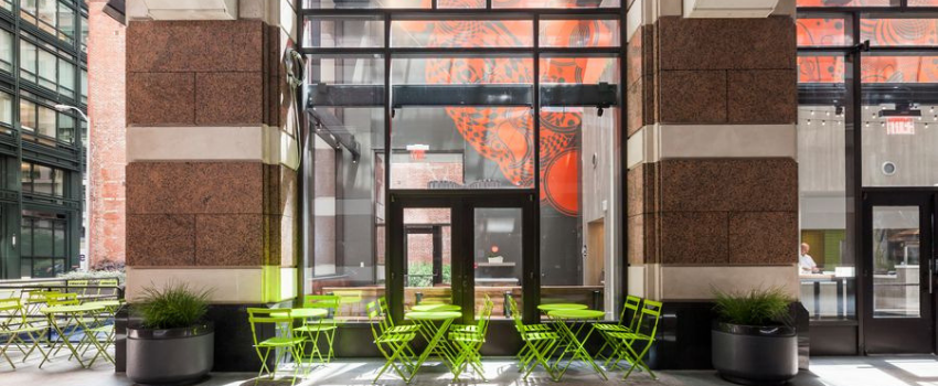 Inside of a building with green chairs and tables next to plants between large brown and tan pillars.